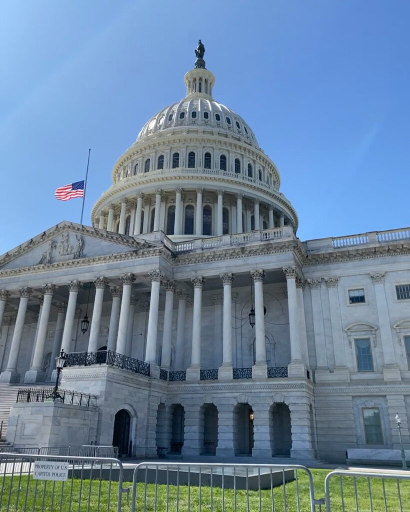 "The United States Capitol building in Washington, D.C., with its iconic white dome and columns, under a clear blue sky. The American flag is flying at half-mast, and metal barricades are set up in front of the building.