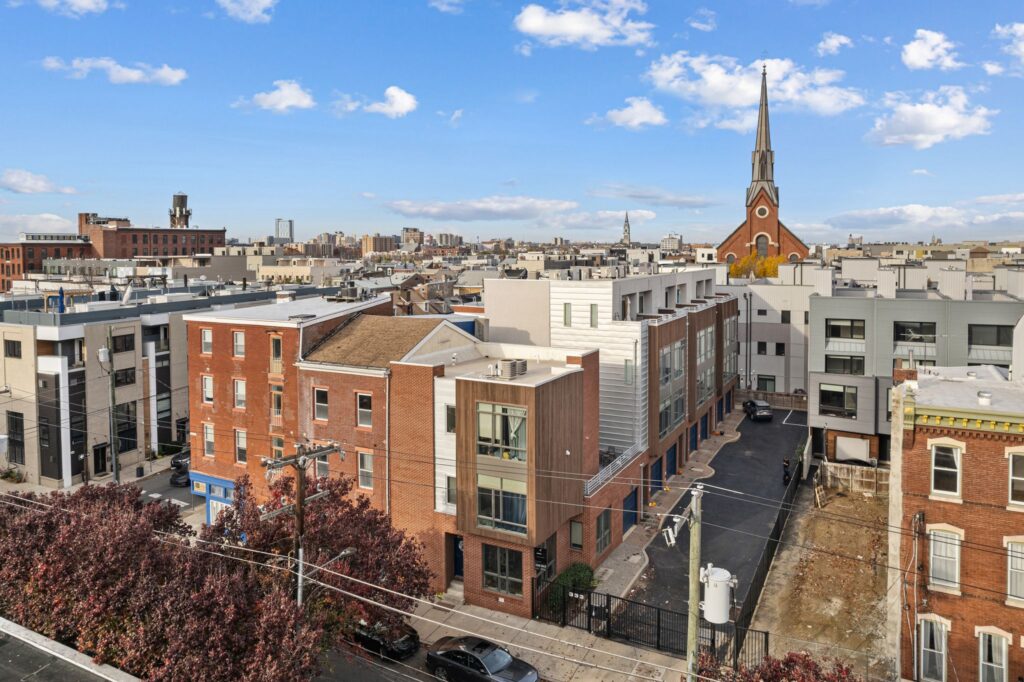 A city block in Northern Liberties, Philadelphia, featuring a mix of historic brick rowhomes, modern townhouses, and mid-rise buildings, with a red-brick church and steeple visible in the background under a blue sky.