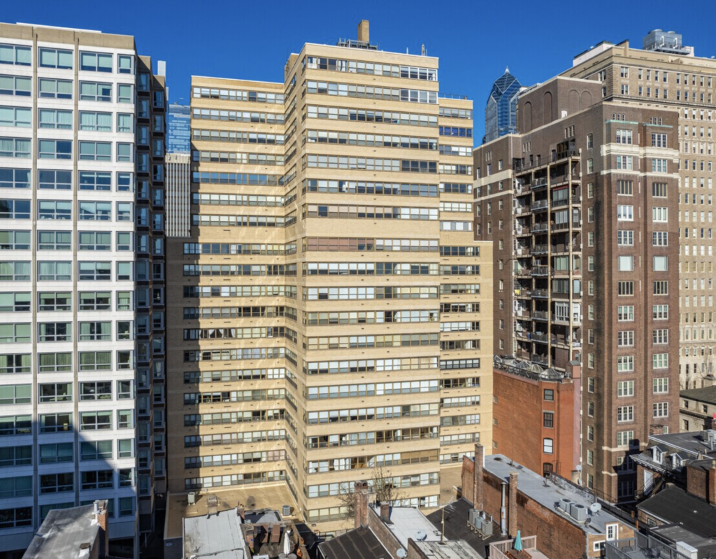 Exterior view of the Rittenhouse Savoy apartment building in Philadelphia, a beige mid-century high-rise with long horizontal rows of windows, surrounded by neighboring residential towers under a clear blue sky.